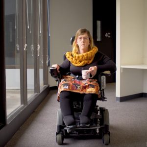A woman in a power wheelchair moves through a hallway holding a patterned mug. She is wearing a chunky mustard-yellow scarf, black top, and a colorful patchwork-style skirt. The hallway has large windows on one side and a door in the background.