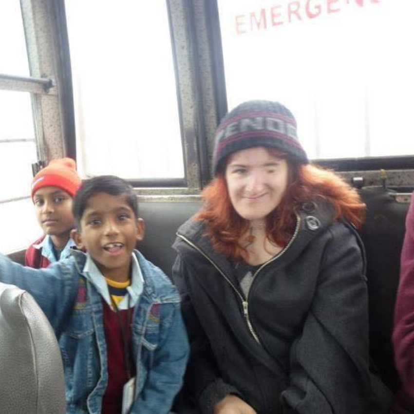 Woman with long reddish hair and wearing a beanie sits at the back of a bus next to two young Indian boys.