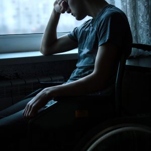 Young man seated in a wheelchair in a dark room by the window. His elbow is resting on the windowsill and his head is resting on his hand. He is wearing a dark t-shirt.