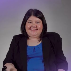 A woman with straight dark hair smiles at the camera while seated in a wheelchair. She is wearing a royal blue blouse under a black blazer, and the background is a neutral grey with soft purple lighting effects in the corners.