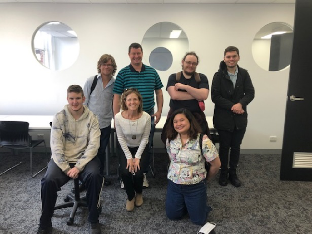 A group of seven people standing together in a meeting room smiling at the camera. Three are kneeling in the front row and four are standing behind them.