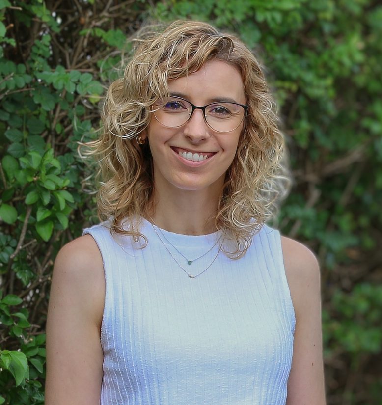 A smiling woman with shoulder length, curly, blonde hair is wearing glasses and a sleeveless white top. There is greenery in the background.
