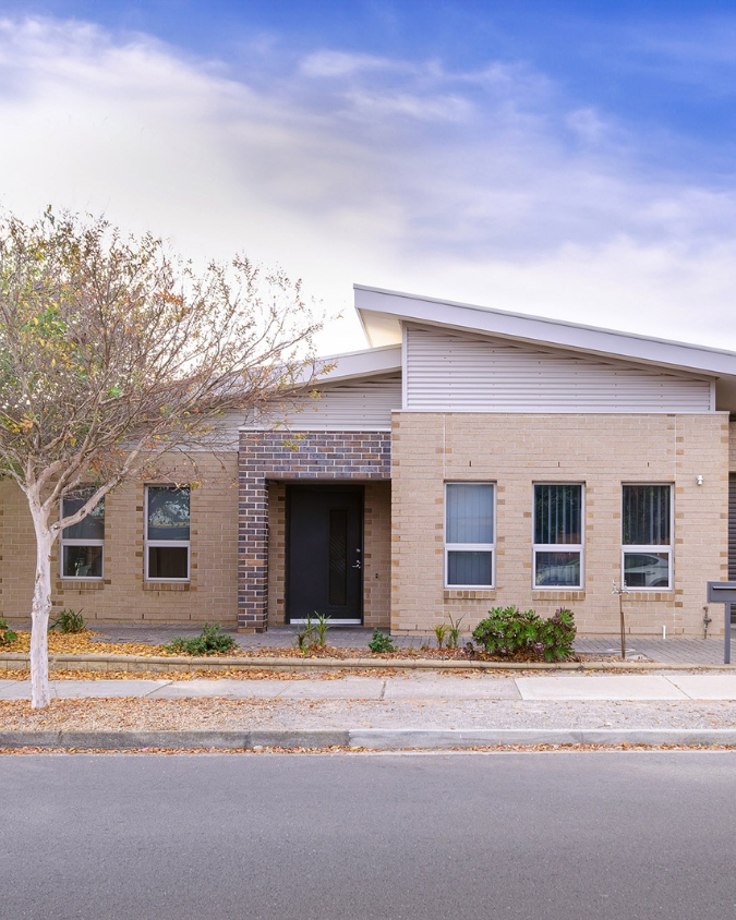 Photo of the front of a light bricked house. It has a sloped roof and a tree to the left. A blue sky with white clouds is visible above the roof line.