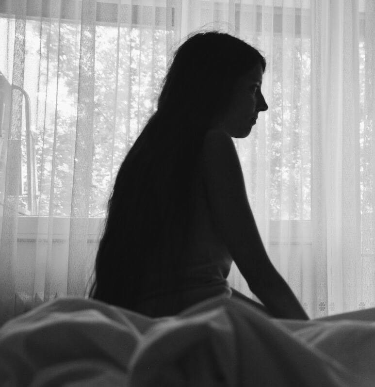 Black and white photo of a woman in shadow sitting on her bed with a window behind her.