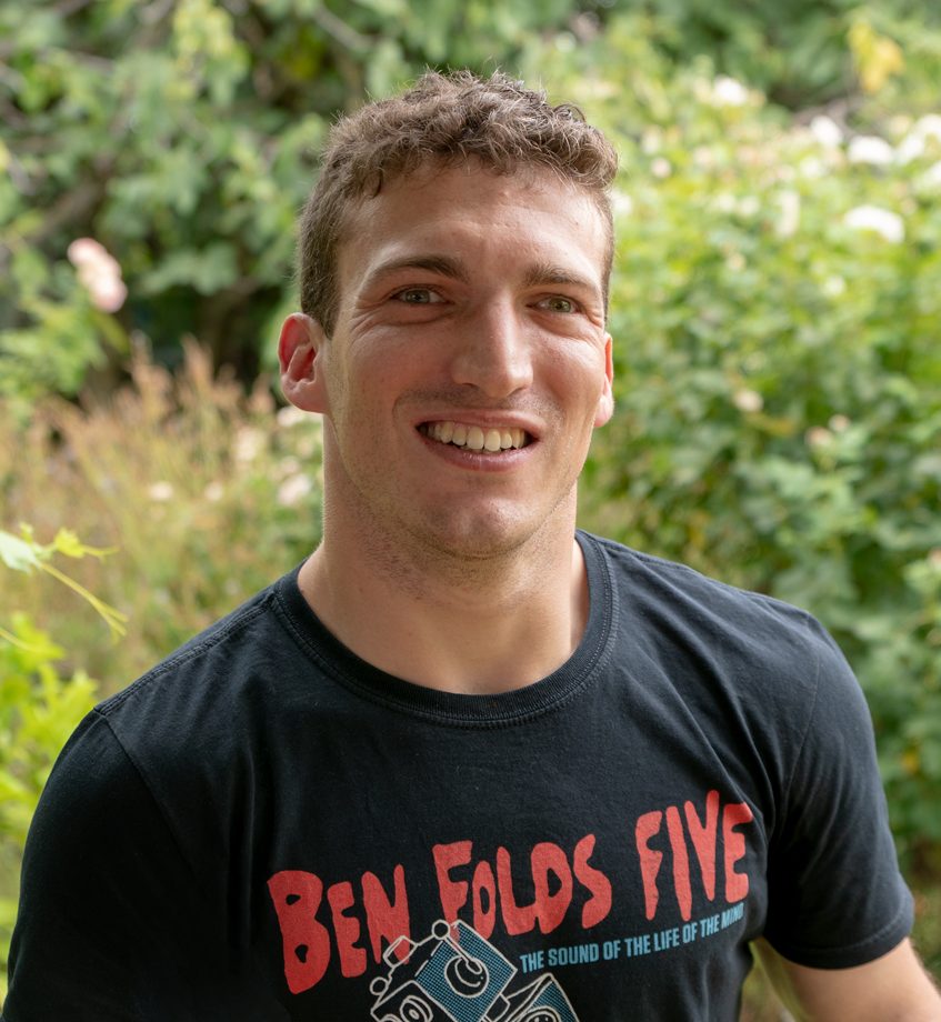 A man with short, wavy brown hair smiles to camera. He is wearing a black "Ben Folds Five" t-shirt with red lettering. The background includes blurred plants and flowers.