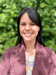Ayla, a woman with long dark hair, is standing in front of blurred greenery and smiling at the camera. She is wearing a brown cardigan over a floral top.