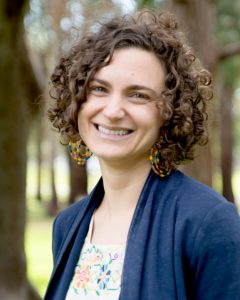 A woman with short, curly hair smiles while standing outdoors. She is wearing large, colorful beaded earrings, a navy blue cardigan, and a light top with a bright, embroidered floral pattern. The background features blurred trees and green grass.