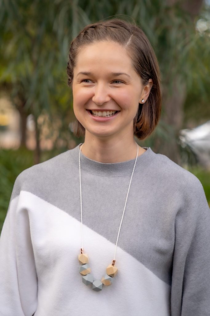 A woman with short, brown hair smiles while standing outdoors. She is wearing a grey and white color-blocked sweater. The background features blurred greenery.