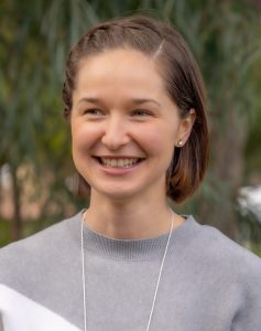 A woman with short, brown hair smiles while standing outdoors. She is wearing a grey and white color-blocked sweater. The background features blurred greenery.