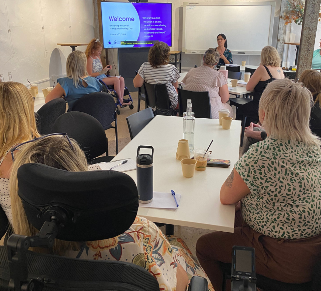 A group of people sit at a table, faced away from the camera towards 2 presenters.