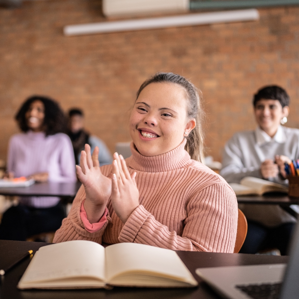 Girl sitting behind a desk with an open book in front of her. She is smiling and has her hands up in front of her as though she is about to clap them. She is looking forward and smiling.