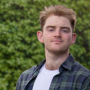 Young man with short reddish hair in a green plaid shirt and white tee, standing before a leafy green background.