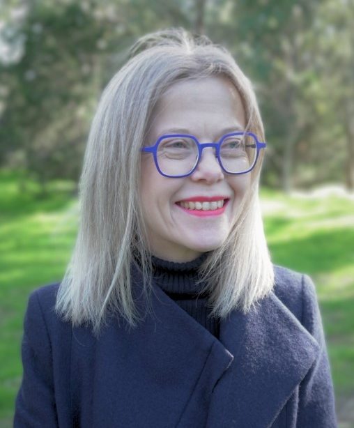 A woman with straight, shoulder-length blonde hair smiles while standing outdoors in a sunlit park. She is wearing bold blue glasses, bright pink lipstick, a black turtleneck, and a dark coat. The background is filled with greenery and softly blurred trees.