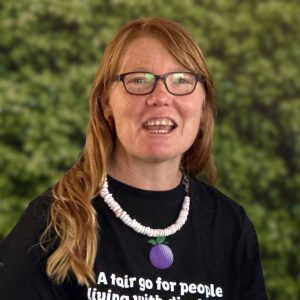A woman with red hair and glasses smiles, wearing a white-beaded necklace and a black Purple Orange t-shirt, against a leafy green background.