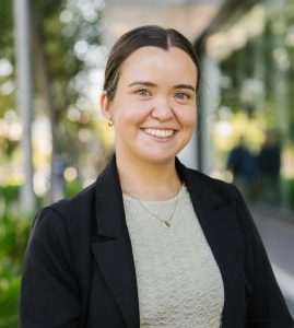 A young woman with dark hair tied back smiles while standing outdoors in a city setting. She is wearing a light textured top, a black blazer, and a gold necklace. Trees and a glass building line the footpath in the background.