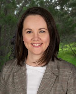 A woman with straight, dark brown hair smiles at the camera while standing outdoors. She is wearing a grey plaid blazer over a white top. The background features softly focused green trees and foliage.