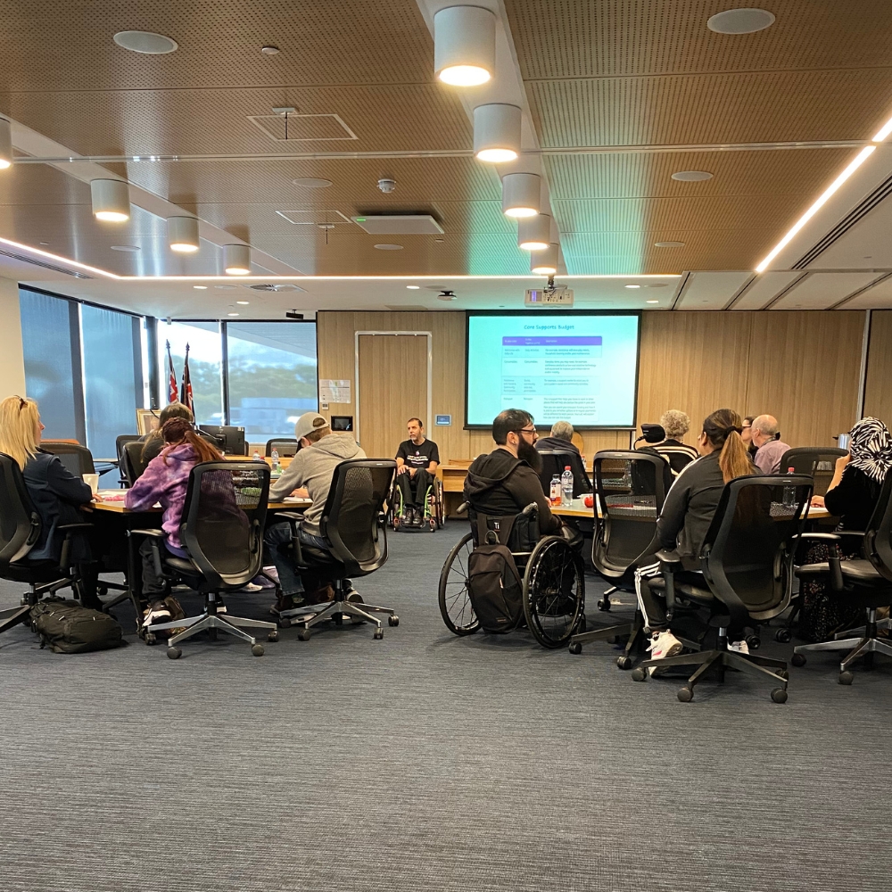 Large meeting room with tables of people and a large presentation screen in the distance.