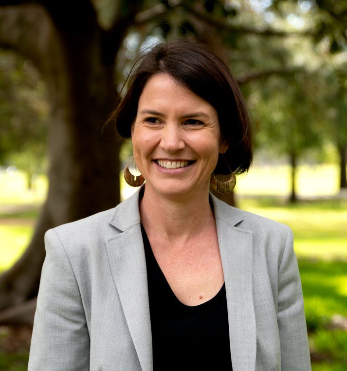 A woman with dark hair smiles while standing outdoors in a sunlit park. She is wearing large circular earrings, a light grey blazer, and a black top. Trees and green grass are visible in the blurred background.
