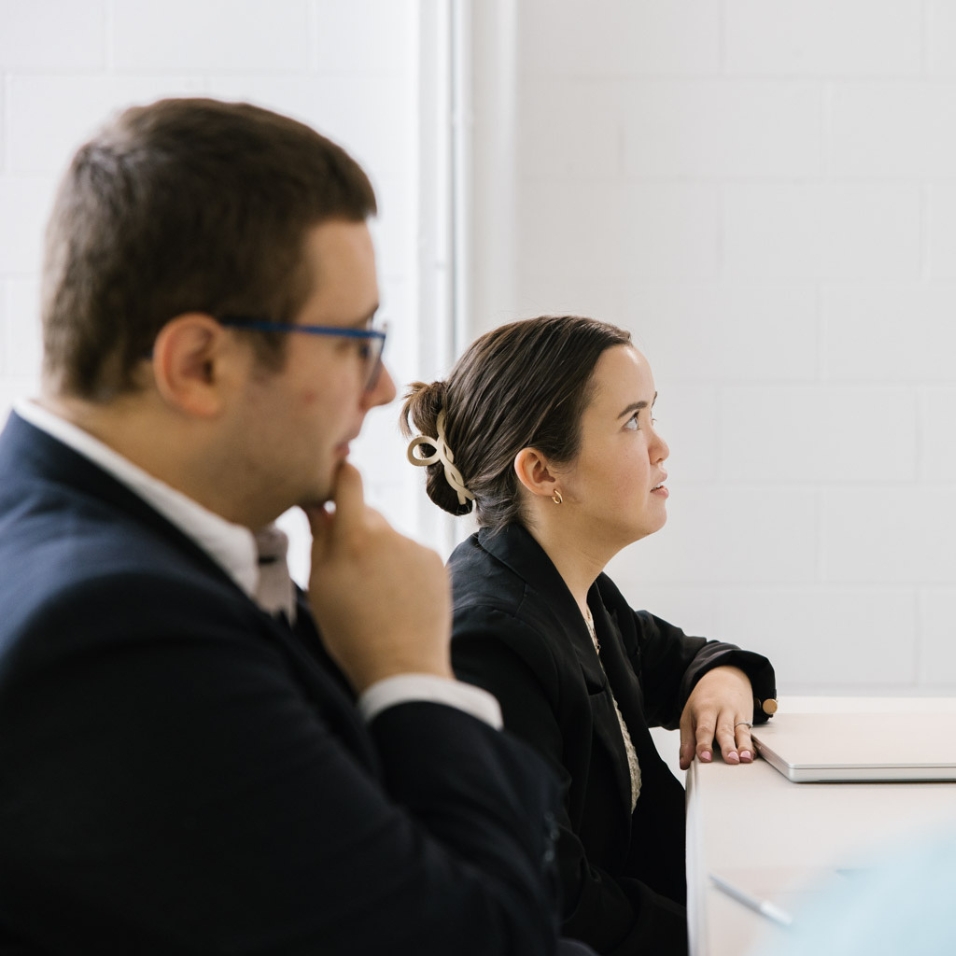 Side view of two people sitting at a desk looking forwards in concentration. They are both wearing dark suits.