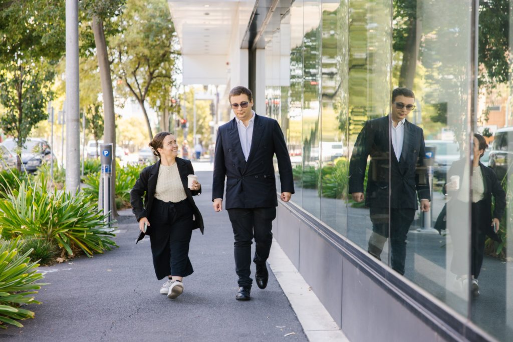 A man and a woman walking along a footpath, talking to one another. Both are dressed in business attire.