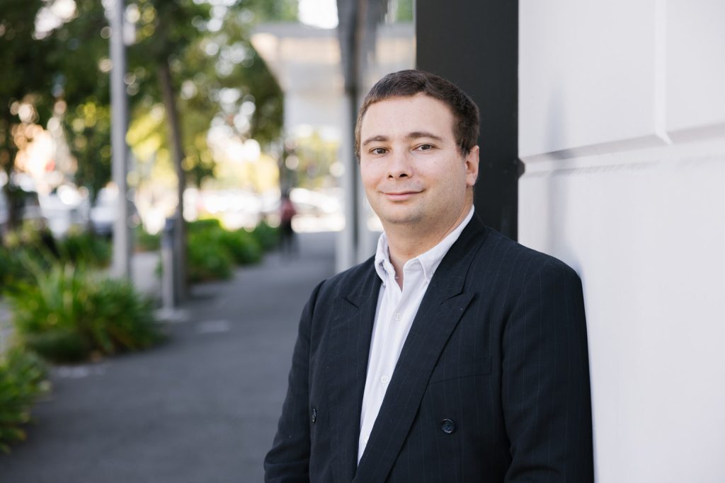 A man with short brown hair is leaning up against a wall. He's wearing a black jacket and white shirt.