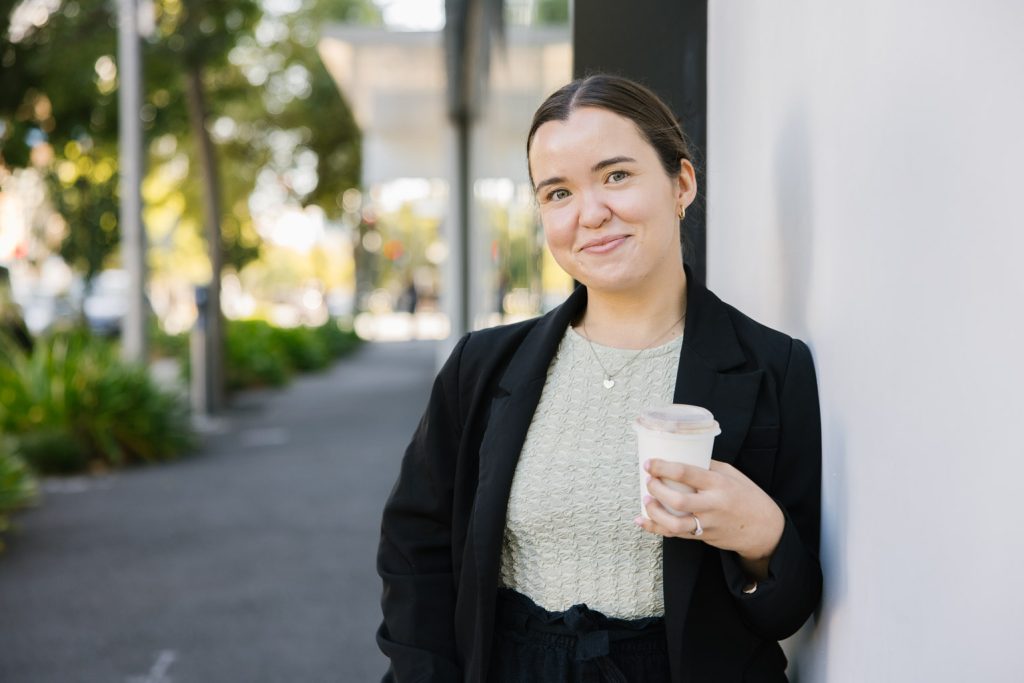 A person with dark brown hair wearing a black jacket and cream-coloured top is leaning up against a wall, holding a cup of coffee.