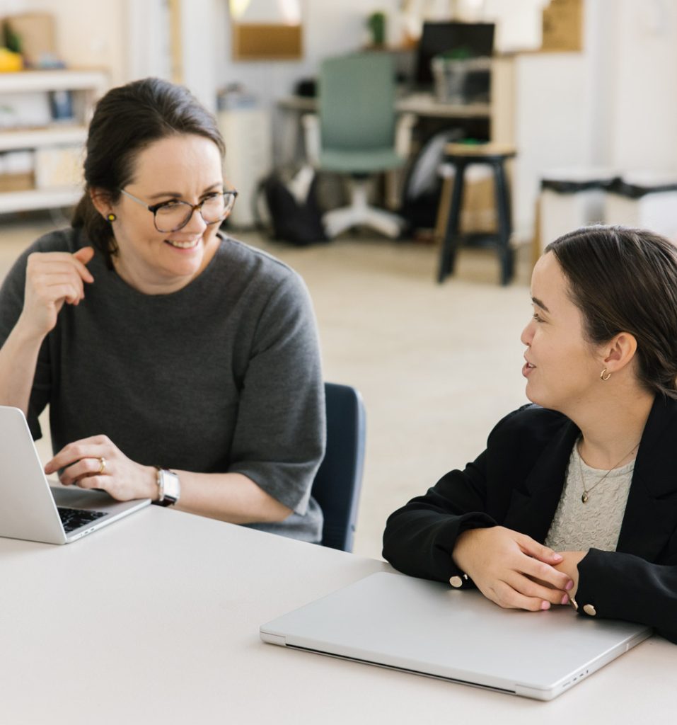 Two women are seated, talking at a table. The woman on the left is wearing a dark grey top and has brown hair and glasses. The person on the right has dark brown hair and is wearing a black jacket and cream coloured top. Both people have laptops in front of them.