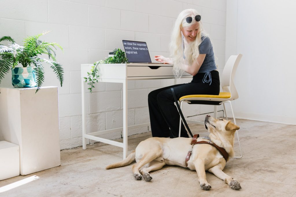 a young woman with long white hair sits at a desk with her assistance dog lying on the floor