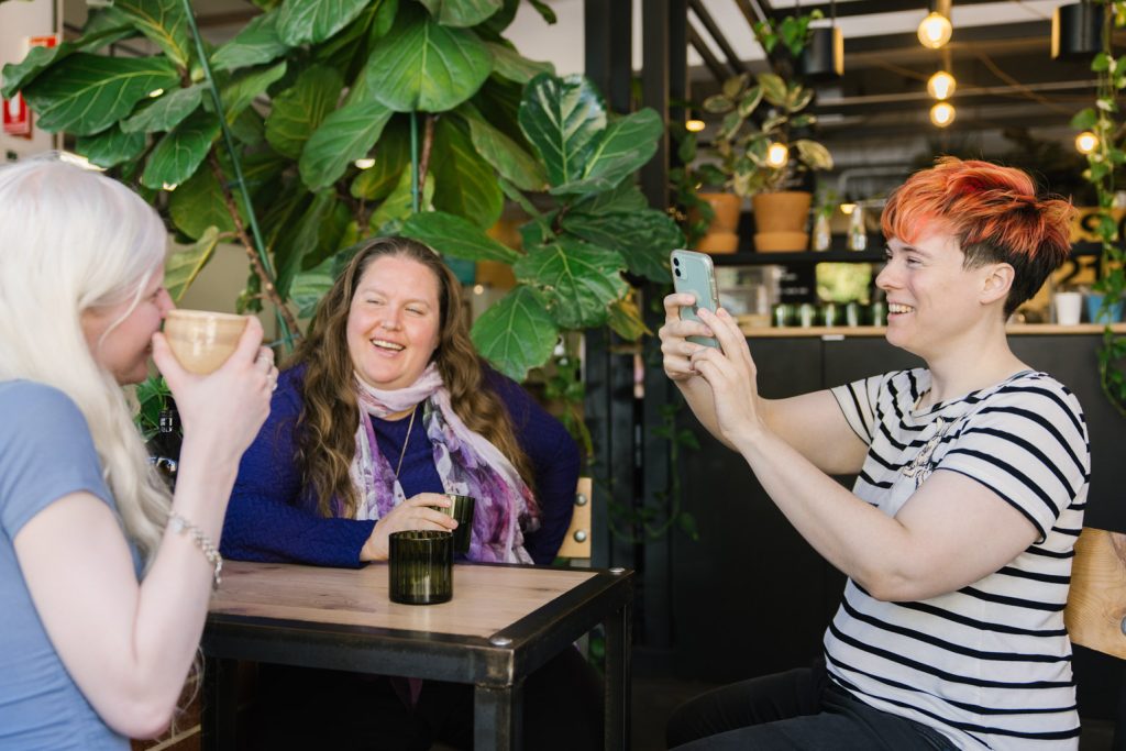 Three people are at a cafe table. One person in at the table is filming another person with a mobile phone, whilst the third person looks on.