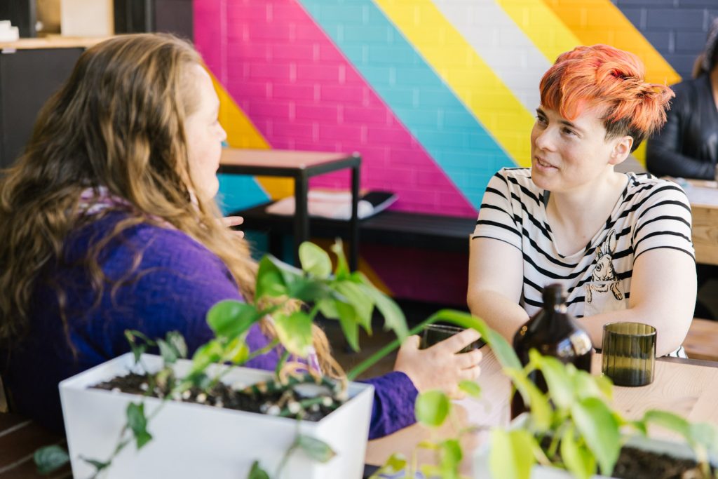 Two people at a cafe table with a rainbow painted wall in the background.
