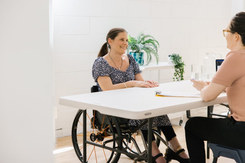 A woman is sitting in a wheelchair at a desk, talking to another person.