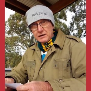 A man wearing glasses and a grey beanie looks toward the camera while holding a piece of paper. He is dressed warmly in a khaki jacket and a green and yellow scarf. Trees and a corrugated metal roof are visible in the background.