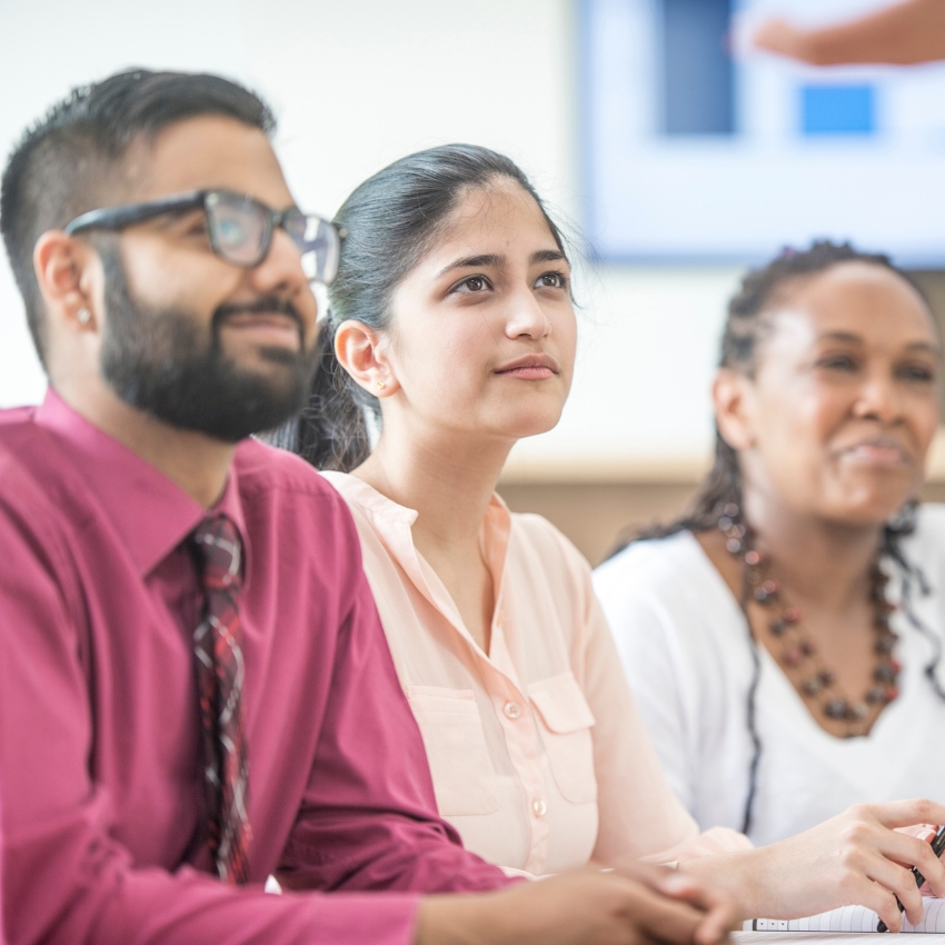 Three people sit looking off camera. They are wearing business attire and seated. There is a man in a red shirt wearing glasses, with two women with dark hair and light coloured tops to the right.