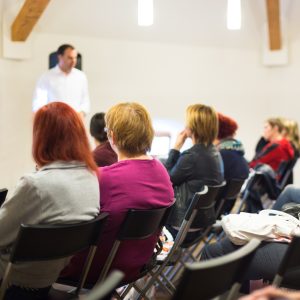 Group of seated people seen from behind. They are in a meeting room and there is a presenter at the front.