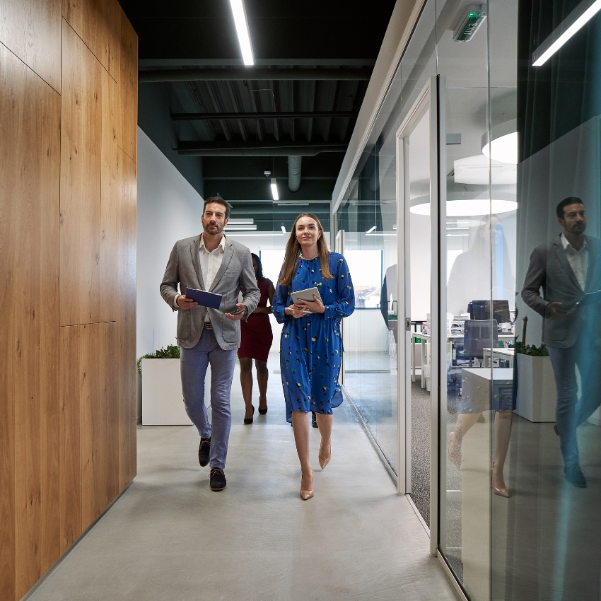 Two people dressed in business attire walking along a corridor in an office building. There is wood paneling on the left and glass office partitions on the right.
