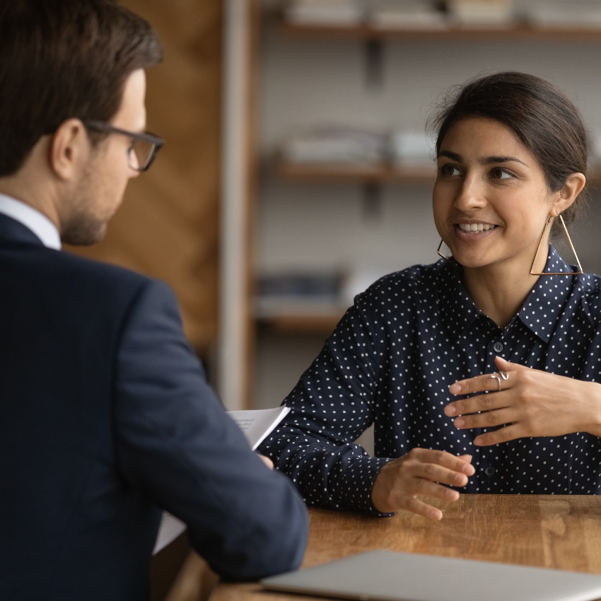 Man and woman seated at a table. The man has dark hair and is wearing a suit - he is pictured from behind looking at the woman on the other side of the table who has dark hair and is wearing a dark shirt. She is smiling and is motioning with her hand.