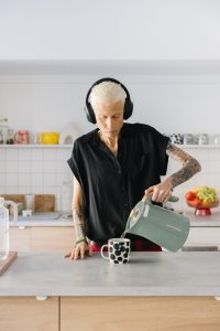 A person stands at a kitchen bench making a cup of tea. They have short bleached hair, tattoos on their arms, and wear a loose black top and headphones.