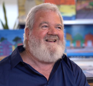 Older gentleman in a dark blue polo shirt and with white hair and beard sits looking off centre and smiling.